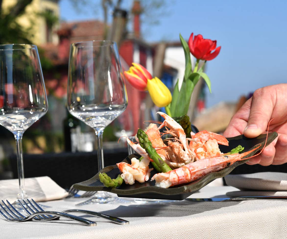 Nel nostro plateatico esterno, in una giornata di sole, una mano serve un piatto di scampi con gli asparagi. Sullo sfondo, il pittoresco canale di Burano che si vede dal nostro ristorante.